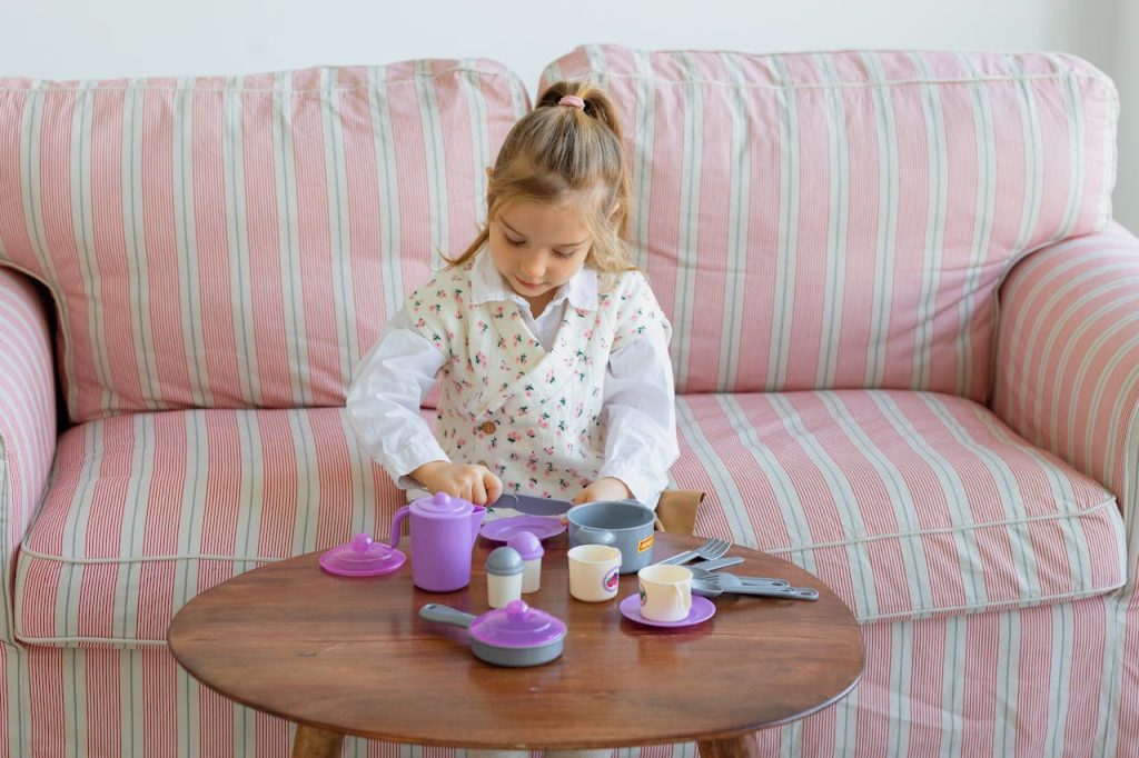 Adorable young girl playing with a toy tea set on a cozy couch in a living room.